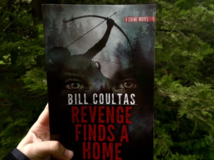 A copy of Revenge Finds a Home is held in front of a dark and grim forest.