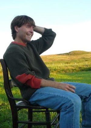 Michael Crummey sits on a wood chair outdoors on a green grassy field. He runs his hand through his hair and laughs.