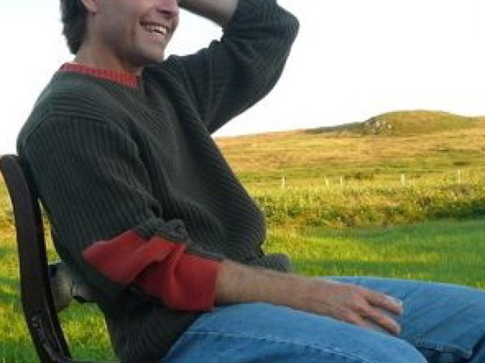 Michael Crummey sits on a wood chair outdoors on a green grassy field. He runs his hand through his hair and laughs.