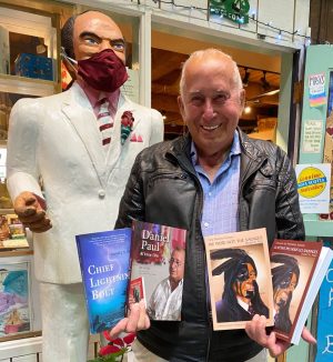 A smiling man stands next to a chainsaw-carved statue of a former prime minister. The man is holding a handfuls of books